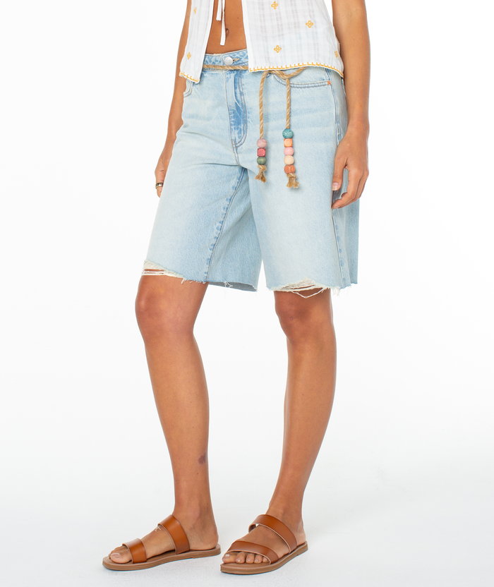 A person wears Roxy's Message In A Bottle Short in light blue with frayed hems, paired with a white patterned shirt, beaded rope belt, and brown sandals. Photo is set against a plain white background.