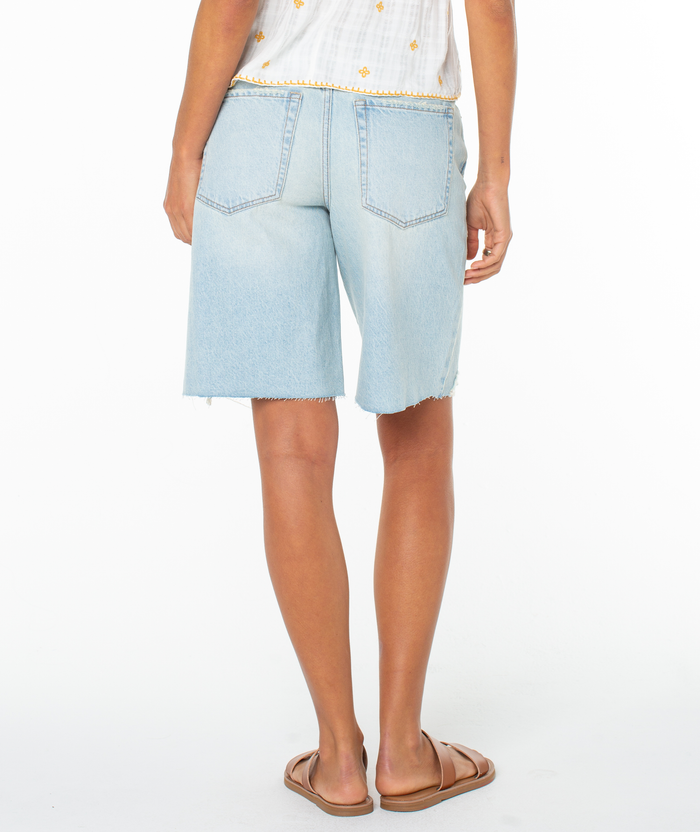 A person stands facing away, wearing the Roxy Message In A Bottle Short with frayed hems, a white patterned top, and brown slide sandals against a plain white background.