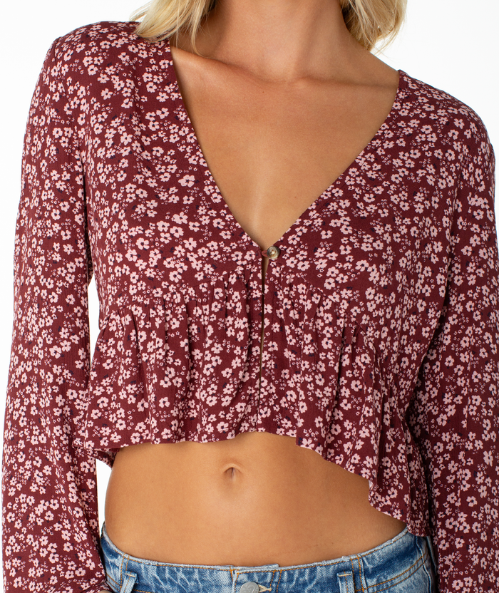 A person wearing the Roxy Night Sky Top—a maroon long-sleeve blouse with a deep V neckline, ruffled hem, small white floral print, and an elegant cropped drape—paired with blue jeans.