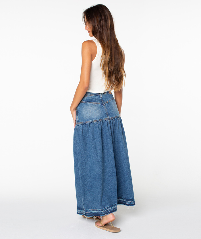 A woman stands sideways against a plain white background, wearing the Roxy Nocturne Prairie Skirt—a long, vintage-inspired denim skirt—paired with a white sleeveless top and sandals. She has long brown hair, hands in her pockets, and is looking down.