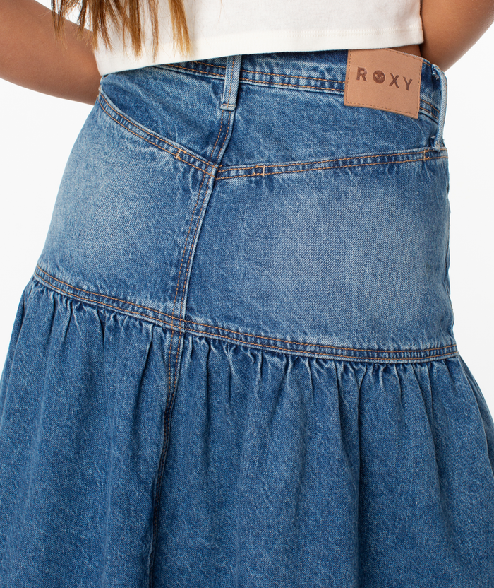 Close-up of a person wearing the Roxy Nocturne Prairie Skirt, a vintage-inspired blue denim skirt with gathered details and a brown ROXY label on the waistband, paired with a white top.