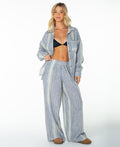 A woman stands smiling in vacation-ready Roxy Offshore Pants in Mood Indigo Coastline Stripe with a button-up shirt, black bra, and brown slippers against a white background.