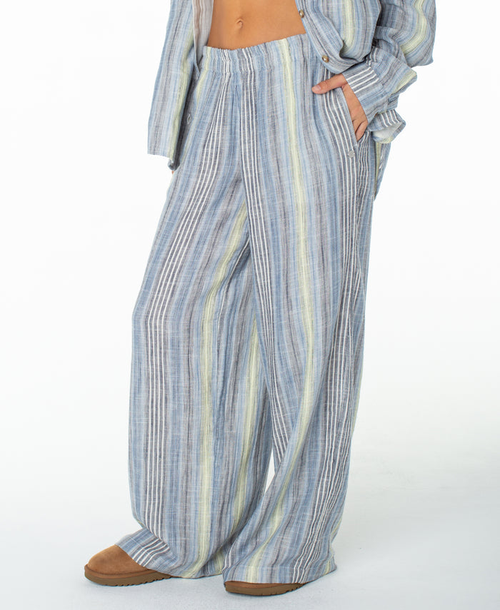 A person is seen wearing the Roxy Offshore Pant—loose, wide-leg pants featuring vertical blue, white, and yellow stripes in soft cotton for a laid-back, vacation-ready vibe—paired with a matching button-up shirt and brown shoes. The upper body is mostly out of frame.