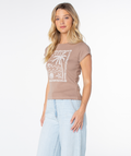 A woman with long blonde hair wears the Roxy Palm Poster 90S Baby Tee in Ginger Snap Palm Poster and light blue jeans, standing against a plain white background and looking forward with a relaxed expression.