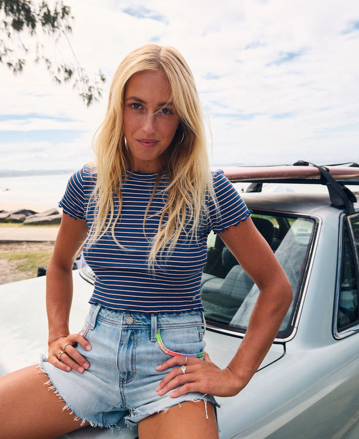 A woman with long blonde hair, dressed in the Roxy Poolside Crop Tee and denim shorts, sits on the hood of a light blue car by the beach, looking at the camera on a bright, sunny day and effortlessly showcasing summery flair.