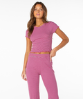 A woman stands against a white background wearing the Roxy Poolside Crop Tee in pink and white stripes, paired with matching pants. One hand rests on her hip while the other points to her pants, creating a perfect fresh summer look.