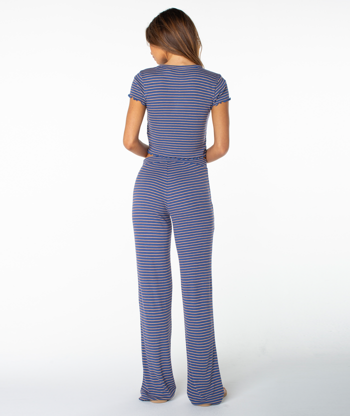 A person stands facing away from the camera against a plain white background, wearing the Roxy Poolside Crop Tee in blue and purple stripes with matching wide-leg pants, infusing summery flair into an everyday wardrobe staple.