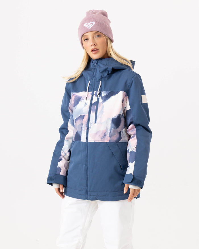 A woman wearing the Roxy Presence Parka Jacket in a blue and white pattern with 15K protection, paired with white pants and a light pink beanie, stands against a plain white background.