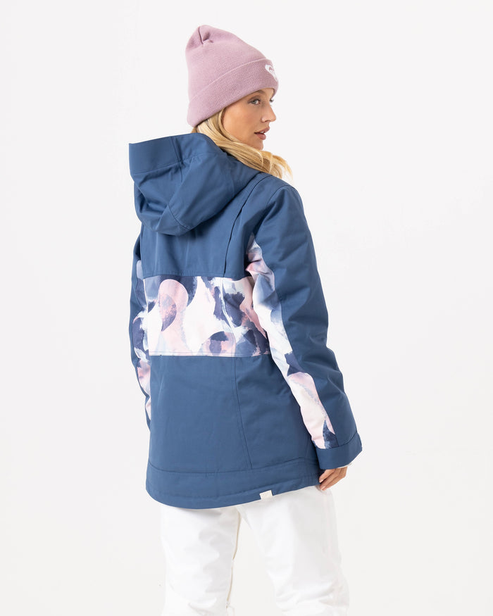 A person is shown from behind, turning slightly to the side against a plain white background, wearing the Roxy Presence Parka Jacket—a women’s snow jacket with 15K protection in blue with a pink and white abstract stripe on the back—paired with white pants and a light purple beanie.