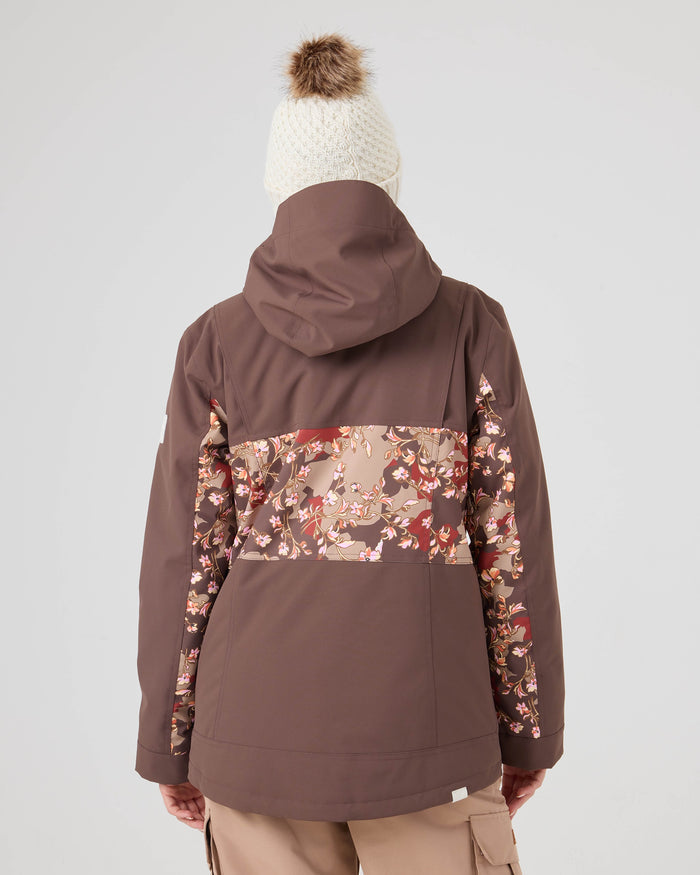A person is seen from behind wearing the Roxy Presence Parka Jacket, a brown women’s snowboard jacket with a floral pattern across the back, paired with a white knit beanie featuring a pom-pom, standing against a plain light background.