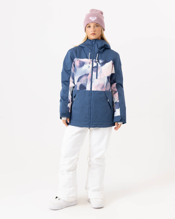 A woman wearing a pink beanie, white snow pants, and the Roxy Presence Parka Jacket in Ensign Blue with 15K protection stands against a plain white background.