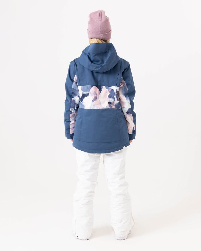 A person stands with their back to the camera, wearing a pink beanie and the Roxy Presence Parka Jacket, which has a blue base with a white and pink abstract pattern across the back, paired with white pants, set against a plain white background.