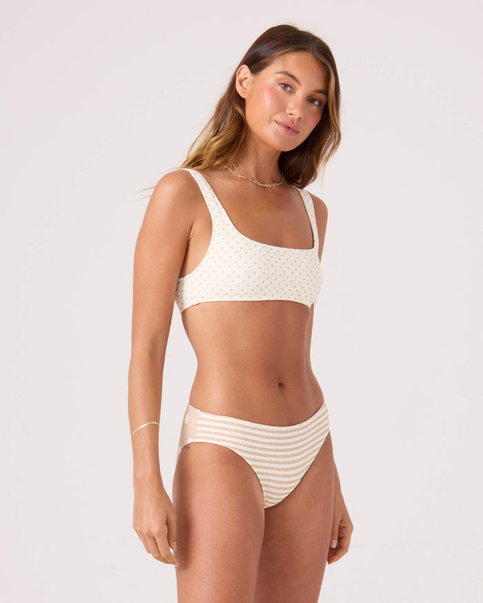 A woman with long brown hair wears the Roxy Polka Dots Square Neck Bralette Bikini Top and high-waisted striped bottoms. She stands against a plain light background, looking at the camera with a slight smile.