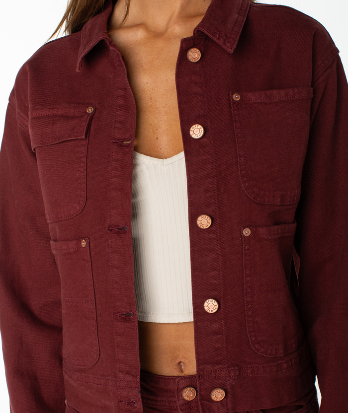 A person wearing the Roxy Reel Life Jacket, a maroon cropped jacket with oversized pockets, layered over a white ribbed crop top, shown from shoulders to hips against a white background.