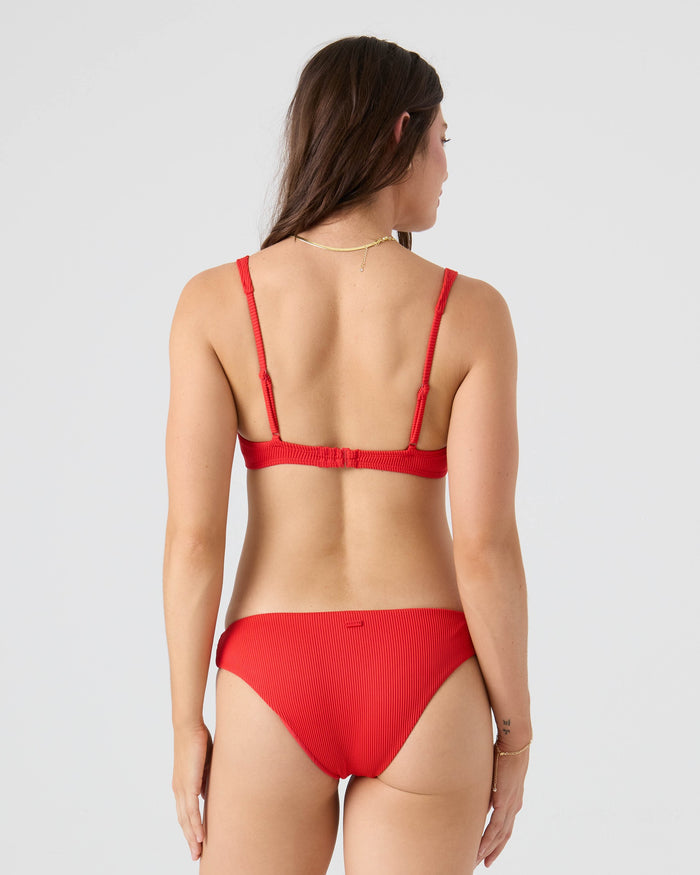 A woman with long brown hair stands with her back to the camera, wearing the Roxy Rib Roxy Love Hipster Moderate Bikini Bottom in red, made from recycled nylon, offering moderate coverage against a plain light background.