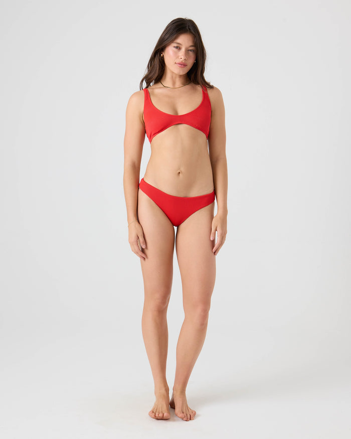 A woman with long brown hair stands barefoot against a plain light gray background, wearing the Roxy Rib Roxy Love Scoop Bralette Bikini Top and matching bottoms made from recycled nylon. She has a neutral expression with arms relaxed at her sides.