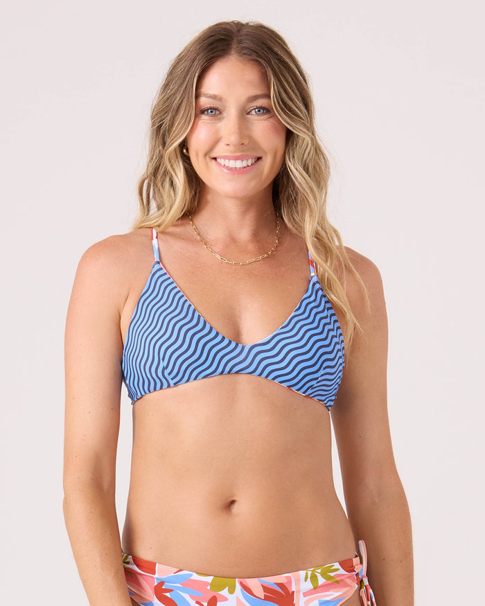 A woman smiles, wearing the Roxy Ripple Effect Reversible Athletic Triangle Bikini Top in Blue Multi with a criss cross back and floral bottoms, posing against a plain light background.