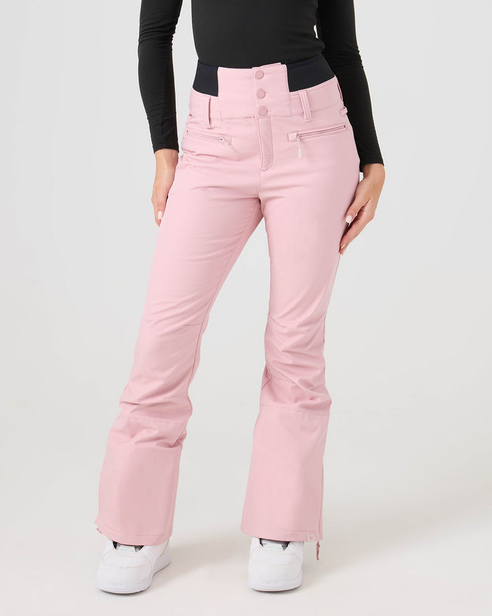 A person wearing the Roxy Rising High Pants in Zephyr—a high-waisted, light pink flare ski pant with side zippers—paired with a black long-sleeve top and white sneakers, standing against a plain light background.