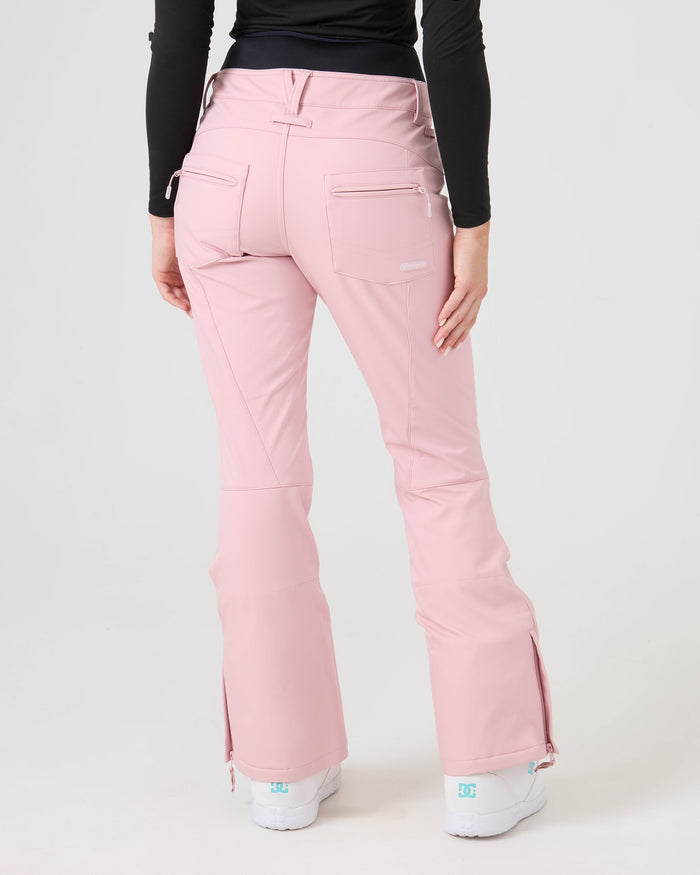 A person is seen from the back wearing Roxy’s Rising High Pants in light pink, featuring back pockets, visible seams, and ankle zippers. They pair the snow pants with a black long-sleeve top and white shoes.