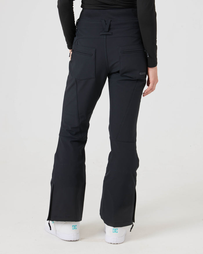 A person is photographed from behind against a plain light background, wearing Roxy’s Rising High Pants—black flared women’s snow pants with back pockets and white sneakers. The softshell fabric provides 15K/15K protection for comfort and durability.