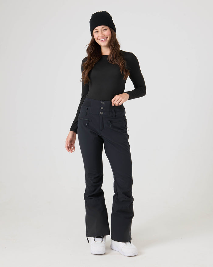 A smiling woman wears a black beanie, a long-sleeve black top, and the Roxy Rising High Pants—high-waisted women’s snow pants crafted from softshell fabric—while standing against a plain white background.