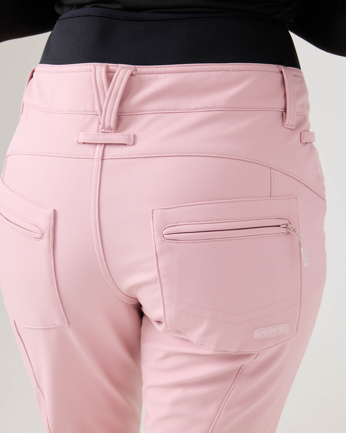 A person wearing the Roxy Rising High Pants in light pink, featuring visible back pockets—including one with a zipper—belt loops, and seam detailing. The pants are styled with a black top, and the photo highlights the waist and hip area from the back.