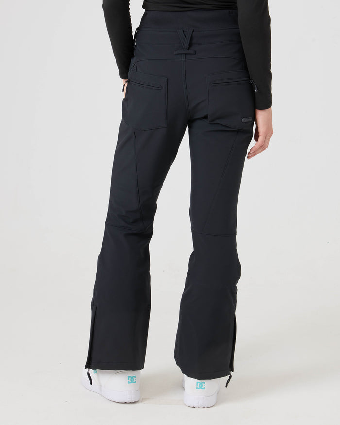 A person is shown from the back wearing Roxy Rising High Pants, a women’s snow pant with 15K/15K protection featuring back pockets and zippered ankle slits. They are also dressed in a black long-sleeve top and white shoes with turquoise accents.