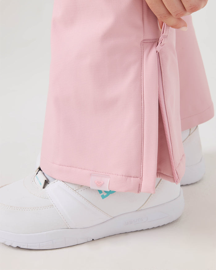 Close-up of a person wearing the Roxy Rising High Pants in light pink, with a side zipper partially open to reveal a white sneaker featuring a blue logo. The person’s hand is holding the zipper.