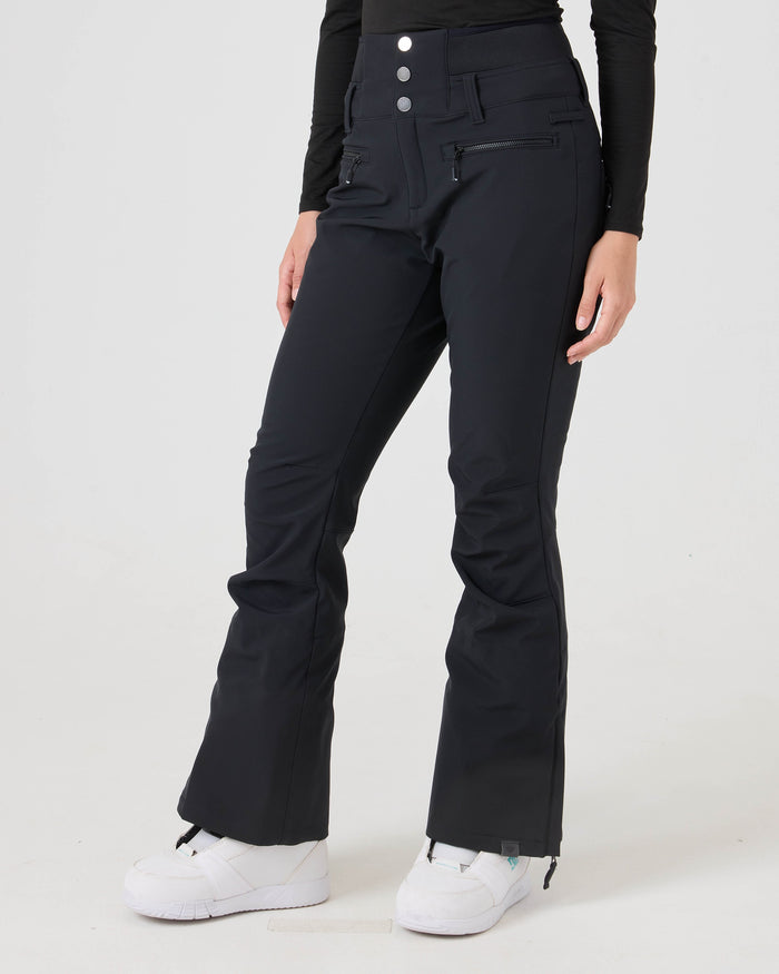 A person wearing Roxy Rising High Pants—a pair of high-waisted black women’s snow pants with front zippers—paired with white sneakers, standing against a plain white background. The softshell fabric provides 15K/15K protection, and only the lower body is visible.
