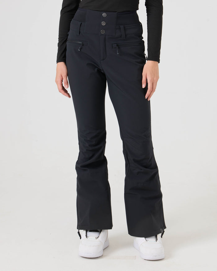 A person is wearing black Roxy Rising High Pants, featuring a high-waisted design and front zippers, along with white shoes, while standing against a plain light background. The image showcases the stylish and functional softshell pants by Roxy from waist to shoes.
