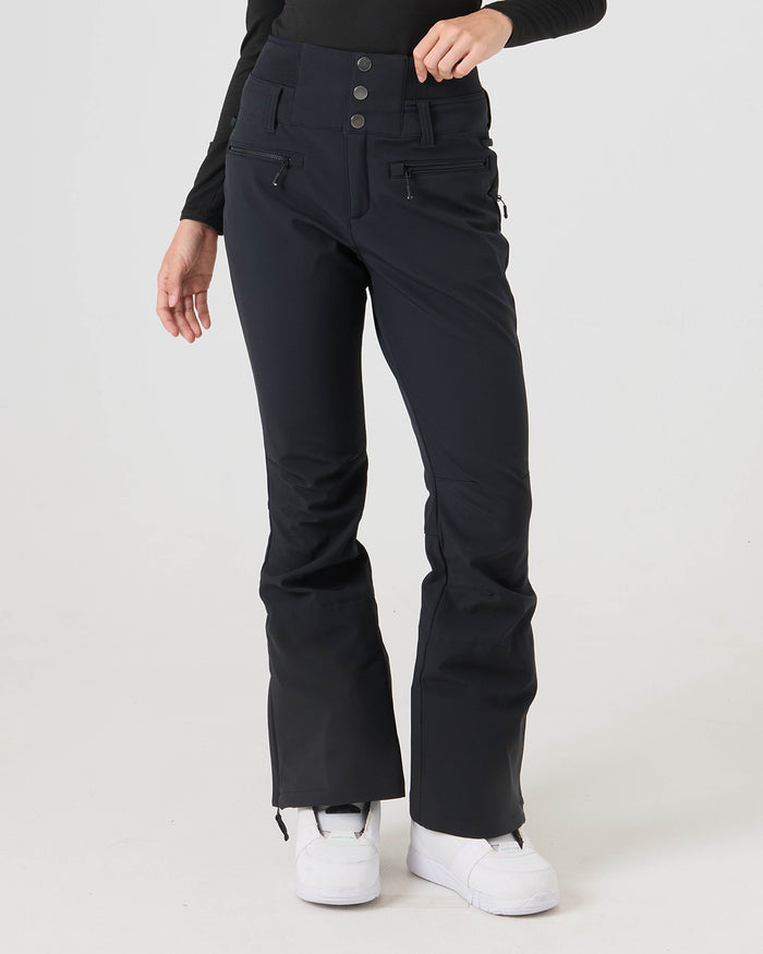 A person wearing the Roxy Rising High Pants—black, high-waisted, flared softshell pants with zippers and buttons—paired with a black top and white shoes stands against a plain white background.