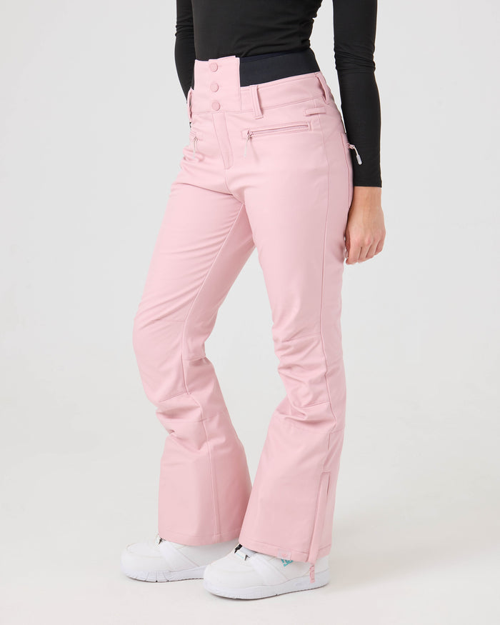 A person wearing Roxy Rising High Pants—high-waisted, light pink flared women’s snow pants featuring zipper pockets and button closures—paired with a black long-sleeve top and white sneakers, stands against a plain white background.