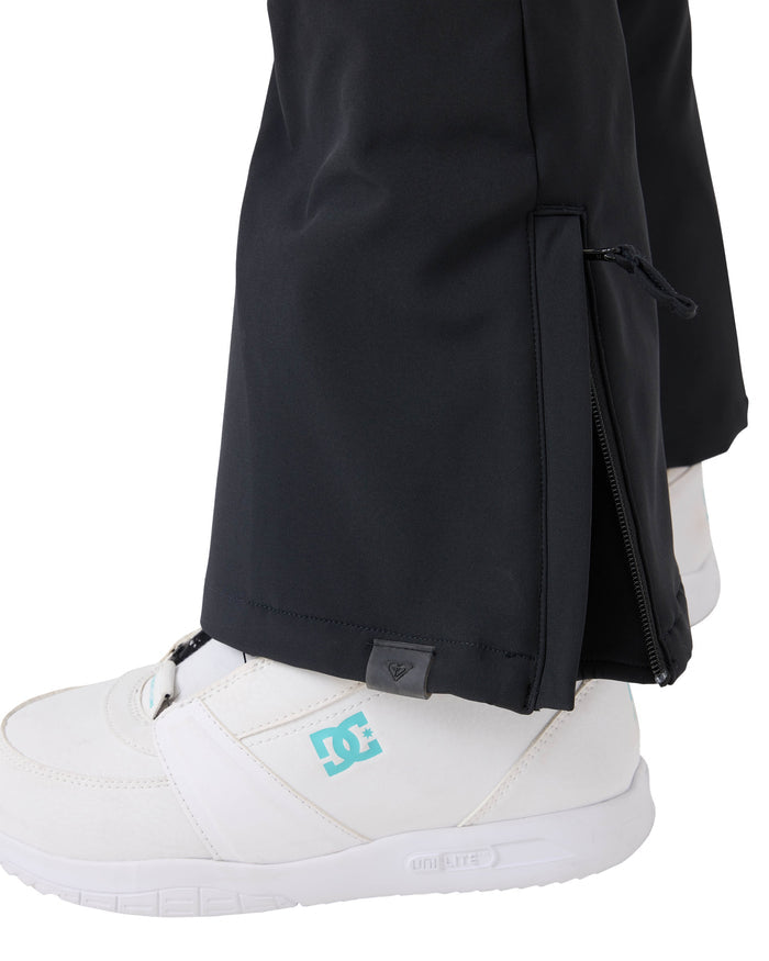 Close-up of a person wearing white DC sneakers and black Roxy Rising High Pants, featuring a zippered leg opening partially unzipped to reveal the shoe underneath. The Rising High Pants, crafted from softshell material for comfort and style, resemble women’s snow pants.