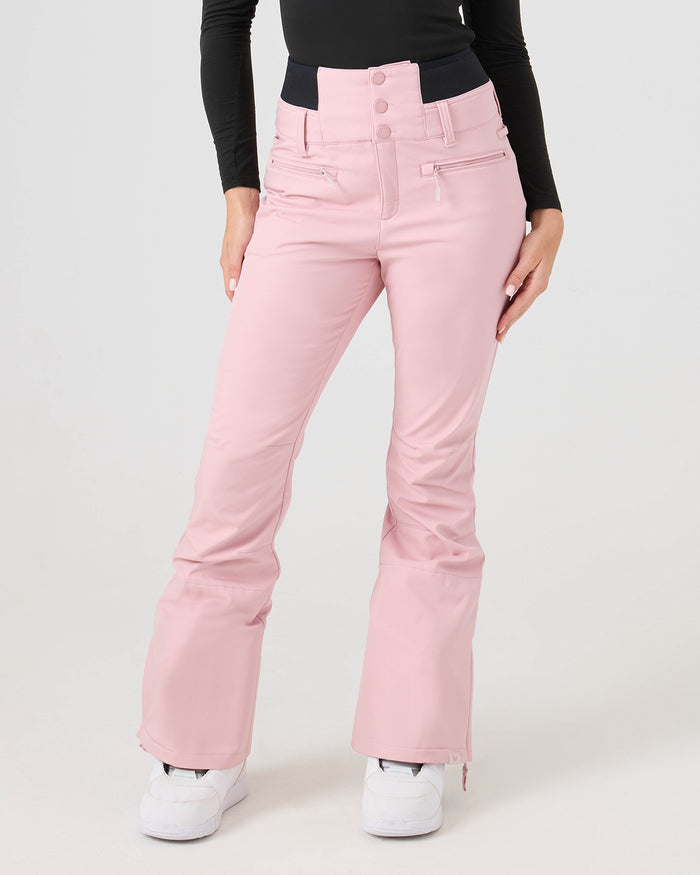 A person wearing the Roxy Rising High Short Pants in Zephyr—a high-waisted, light pink women’s snow pant made from softshell performance recycled fabric—paired with a black long-sleeve top and white sneakers, stands against a plain background.