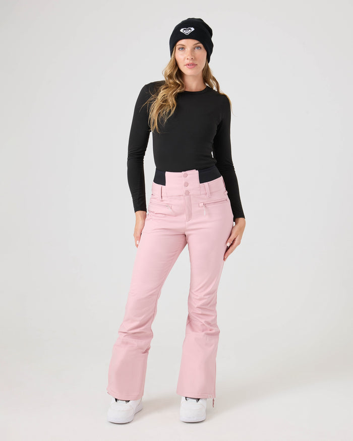 A woman wearing a black long-sleeve top, white shoes, a black beanie, and Roxy’s Rising High Short Pants—pink high-waisted women’s snow pants crafted from recycled fabric—stands against a plain light background looking at the camera.