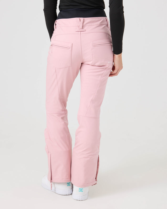 A person is shown from the back wearing Roxy’s Rising High Short Pants, crafted from recycled fabric in light pink. The women’s snow pants feature back pockets and zippers near the ankles, paired with a black long-sleeve top. White shoes with teal accents complete the look against a plain white background.