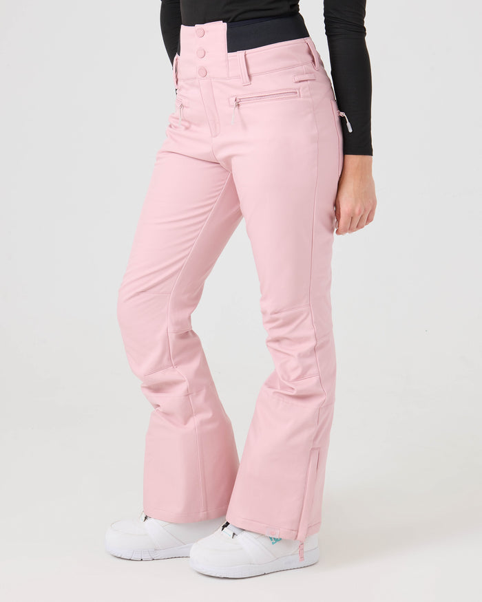 A person is wearing the Roxy Rising High Short Pants, a pair of light pink women’s snow pants crafted from recycled fabric, featuring a high waist, zippered pockets, ankle zippers, and softshell performance for both style and functionality. The pants are paired with a black long-sleeve top and white shoes, set against a plain white background.
