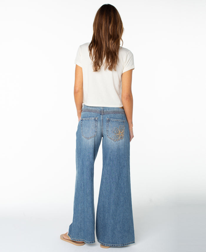 A woman with long brown hair stands facing away from the camera against a white background, wearing the Roxy Rose Heart Cropped Baby Tee—a white short-sleeve top—paired with wide-leg blue jeans for an effortlessly stylish look.