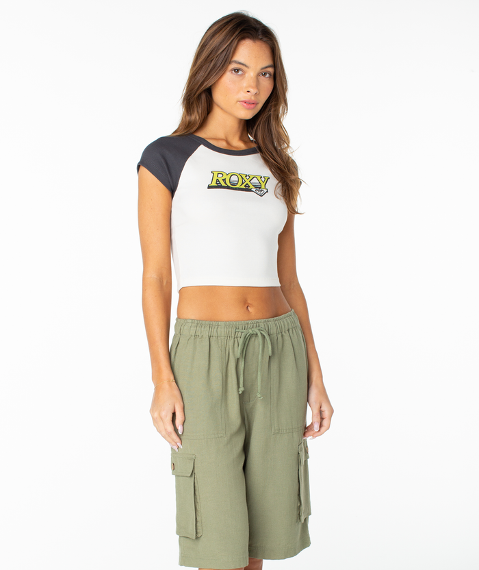 A woman stands against a white background wearing the Roxy Crop Baby Tee paired with olive green cargo shorts—an effortless pick from Roxy for a versatile capsule wardrobe.