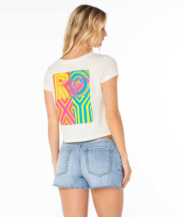 A woman with blonde hair wears the Roxy Echo Tee, a cream-colored T-shirt with a playful graphic and "ROXY" on the back, paired with light blue denim shorts. She stands facing away—a perfect casual summer look.