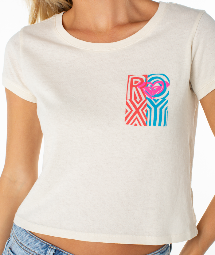 A woman wears the Roxy Echo Tee, a short-sleeved, off-white t-shirt featuring a playful graphic ROXY logo on the left chest, paired with light blue jeans. Her face is not visible.