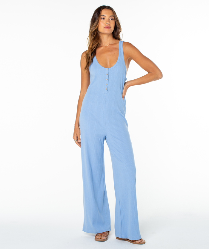 A woman stands against a white background, wearing the Roxy Running Tide Jumpsuit—a sleeveless, light blue piece with front buttons and wide legs—paired with sandals.