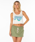 A woman with wavy blonde hair models the Roxy Scoop Baby Rib Tank, a white, cropped cotton top with a colorful logo, paired with a light green drawstring skirt against a plain white background.