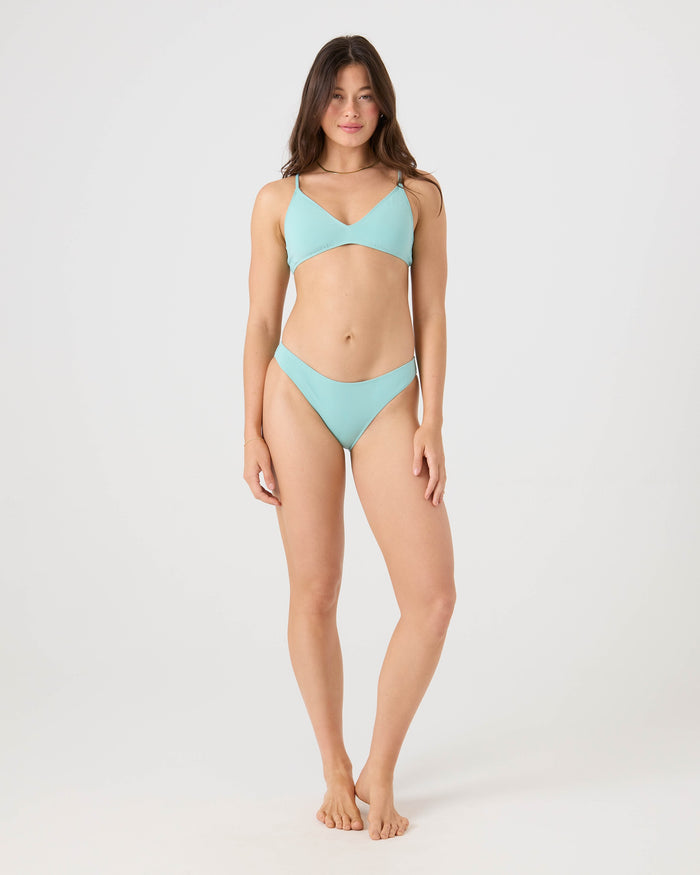 A woman stands barefoot against a white background, wearing the Roxy SD Beach Classics Short Sleeve Athletic Triangle Bikini Top in light blue. She faces forward with arms at her sides and a slight smile.