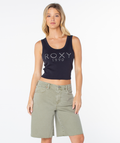 A woman stands against a plain white background wearing the Roxy Shine Bright Scoop Baby Rib Tank in Phantom Shine Bright—a fitted, retro-inspired black sleeveless crop top—and light green knee-length shorts.