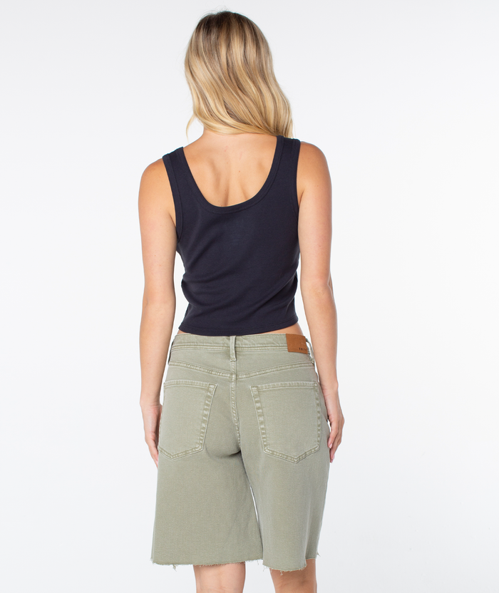 A woman with long blonde hair, wearing the Roxy Shine Bright Scoop Baby Rib Tank and olive green knee-length denim shorts, stands facing away from the camera against a plain white background.