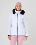 A woman wearing a light pink beanie, light pink pants, and a white Roxy Snowdrift Jacket—a women’s ski jacket featuring a black faux-down fur collar—stands against a plain light background, gazing slightly to her right.