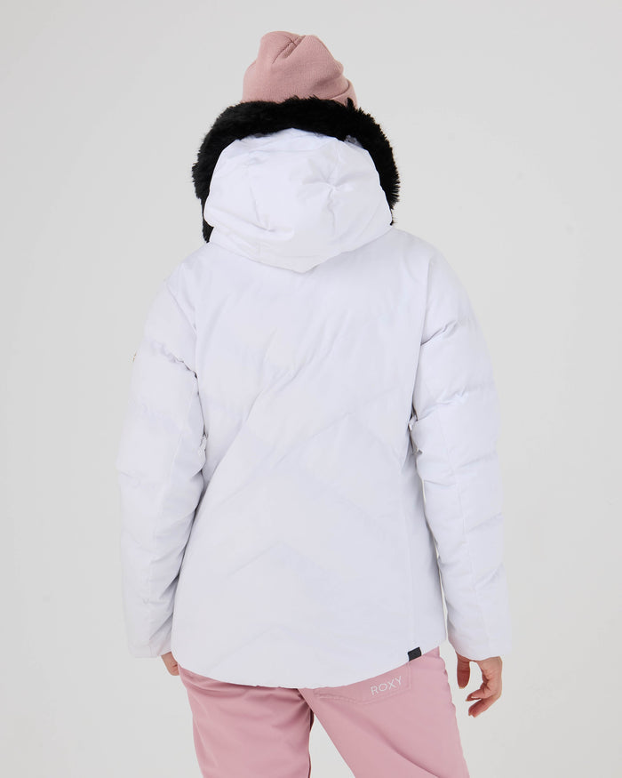 A woman is shown from the back against a plain light background, wearing a white Roxy Snowdrift Jacket—accented with black faux-down fur trim—paired with pink pants and a pink beanie.