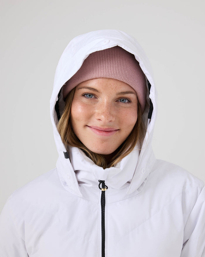 A person in a light pink beanie and the Roxy Snowdrift Jacket—a women’s ski jacket featuring faux-down insulation—smiles at the camera against a plain background.
