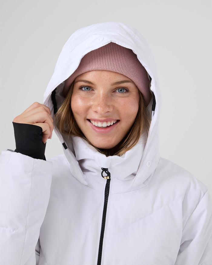 A smiling person wearing the Roxy Snowdrift Jacket, a stylish women’s ski jacket, pulls up the hood and looks at the camera against a plain background.